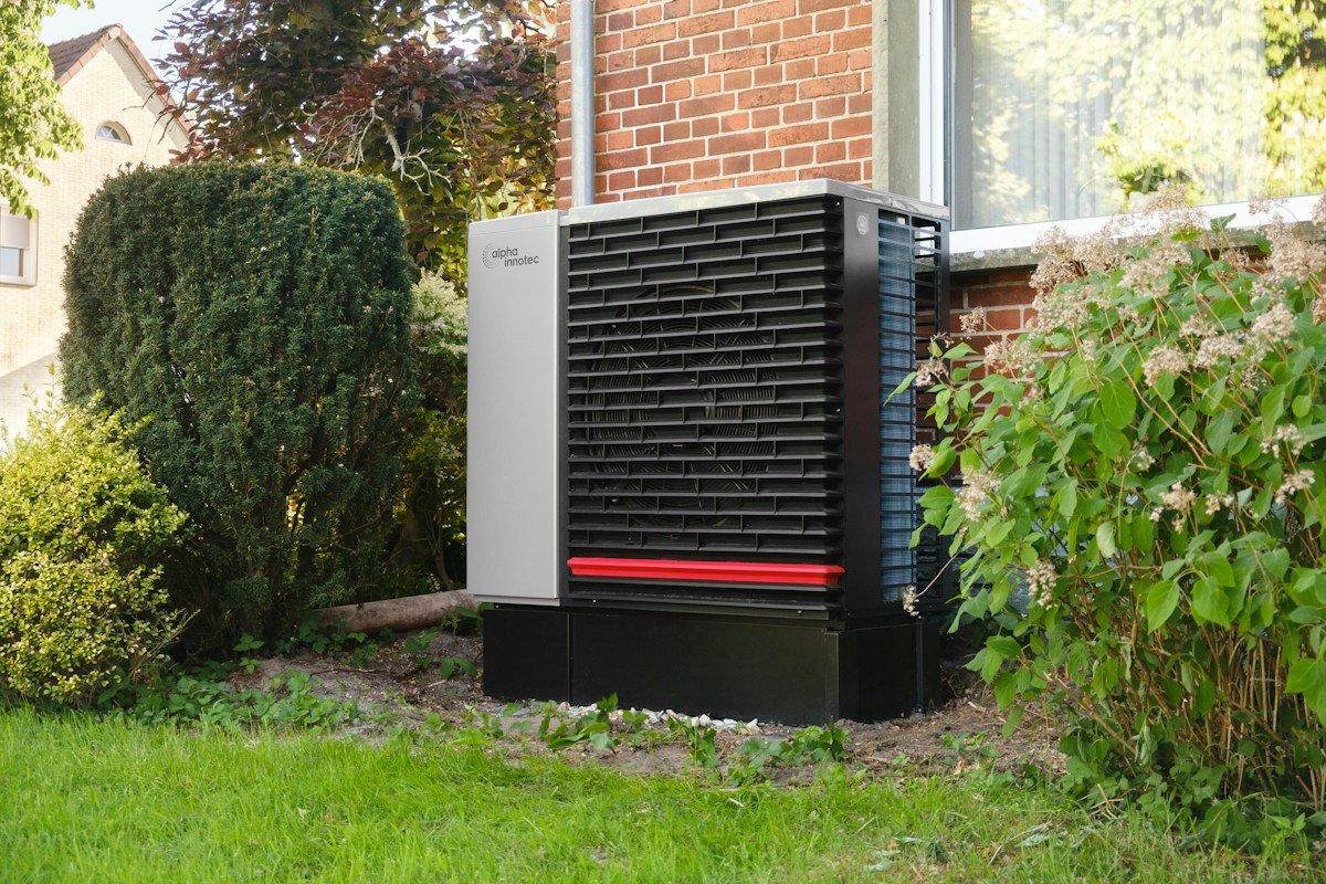 Exterior heat pump unit beside a brick home and garden planting